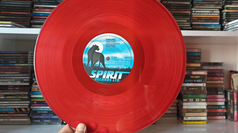 A hand holds a vibrant red vinyl record, with a record collection blurred in the background, creating a nostalgic and music-themed image.
