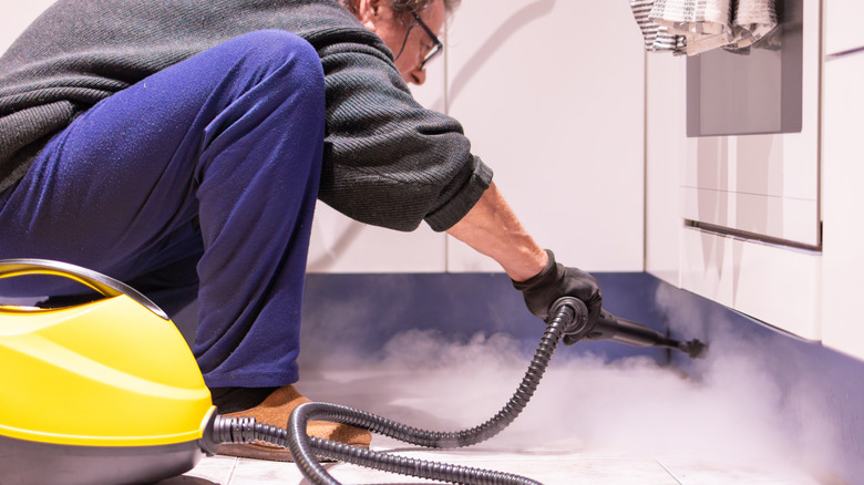 A man is crouched down on a kitchen floor, using a steamer to clean kitchen walls below appliances.