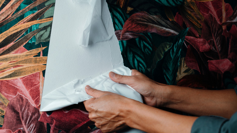 A man's hands pull tropical-patterned wallpaper from a wall.