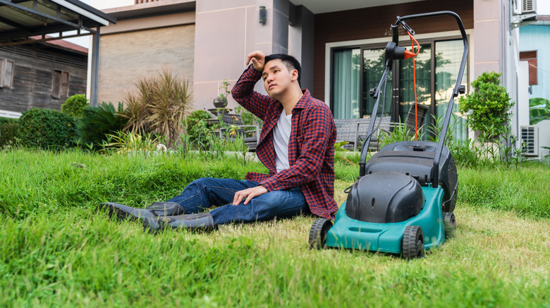 tired man sitting in grass next to lawn mower