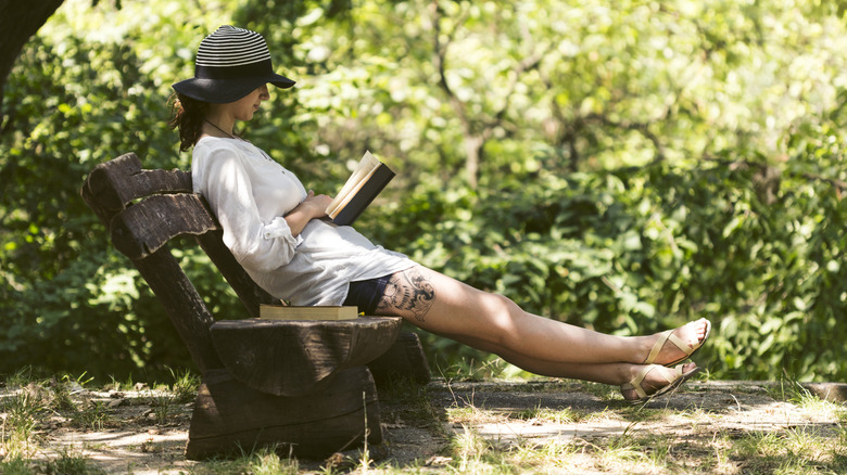 A person reads a book on a bench shaded from the spring sunlight by trees.