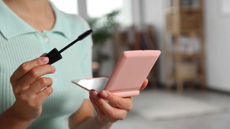 Woman holding a mascara brush in one hand a compact mirror in the other