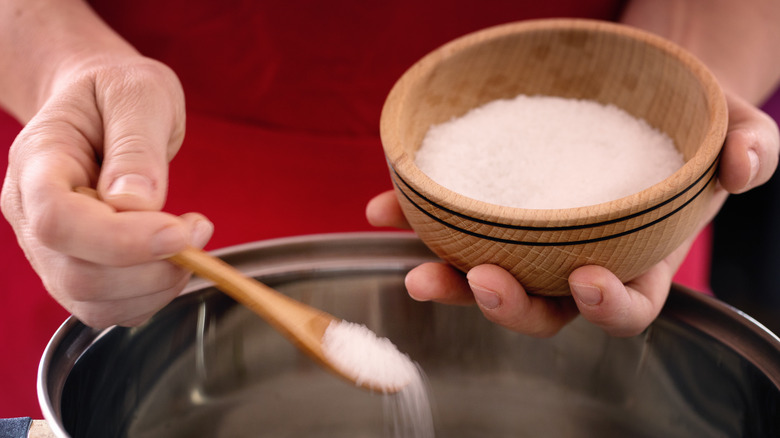Hands holding a bowl of coarse salt and spooning it into pot.