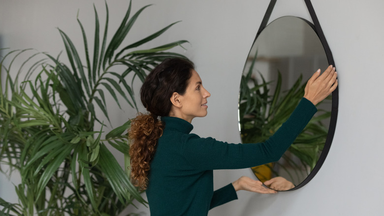 A woman hanging a round mirror next to a plant.