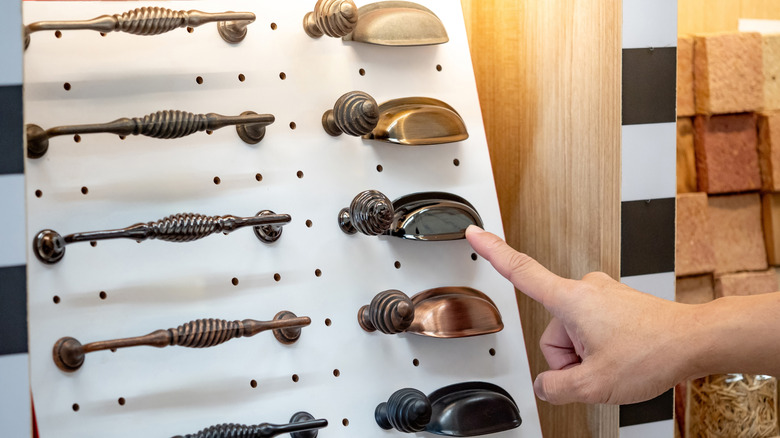 A person points to a dark finish on a cup handle and knob among other finishes and hardware shapes