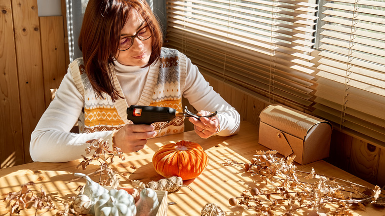 A woman holds a hot glue gun to a piece of decor while DIY fall ornaments.