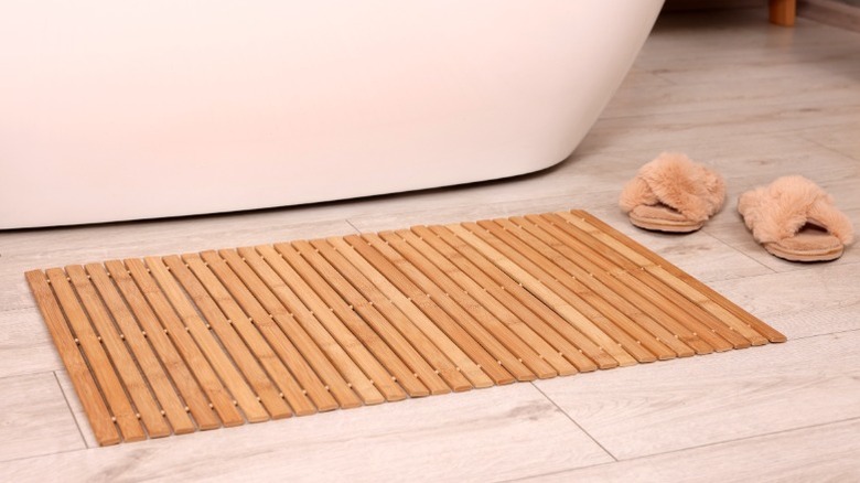 Bamboo bath mat sitting on tile bathroom floor next to slippers and white tub