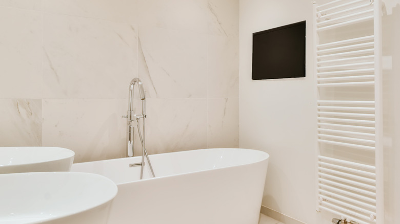 A white marble bathroom featuring a free standing tub with a black TV mounted on the wall