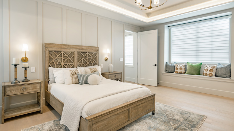 A large bedroom with a queen sized wooden bed, wood flooring, a window, and cushions arranged in a small a window nook