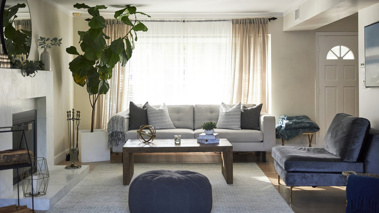 A living room with floor to ceiling drapes, comfortable furniture pieces, a tall plant, and fireplace with a mirror on the wall