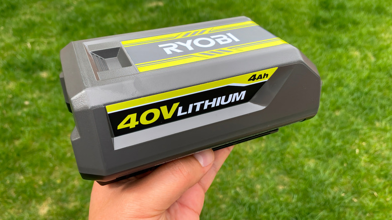 A person holds a legitimate Ryobi power tool battery outside over a lawn.