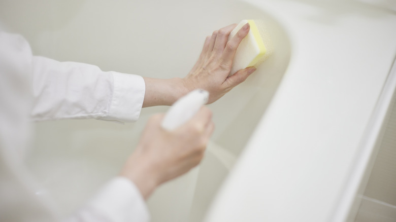 Person with spray bottle and sponge scrubbing a bathtub