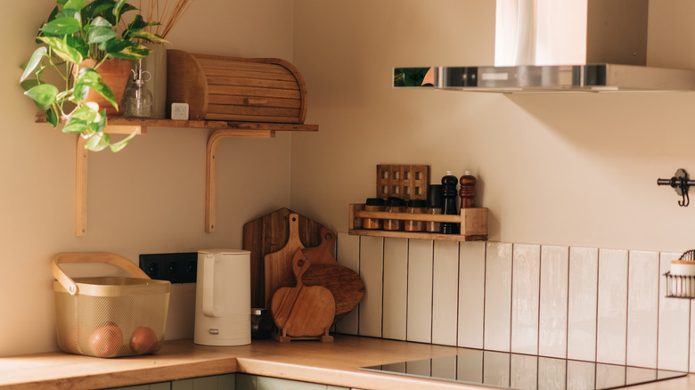 A kitchen counter with stovetop, cutting boards, spice rack, bread box, fruit basket, and hanging plant