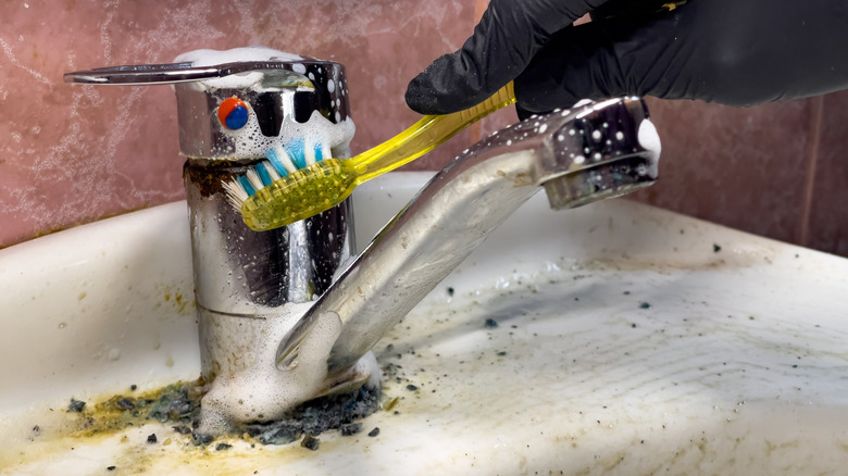 A person cleans a grimy sink faucet with a yellow toothbrush