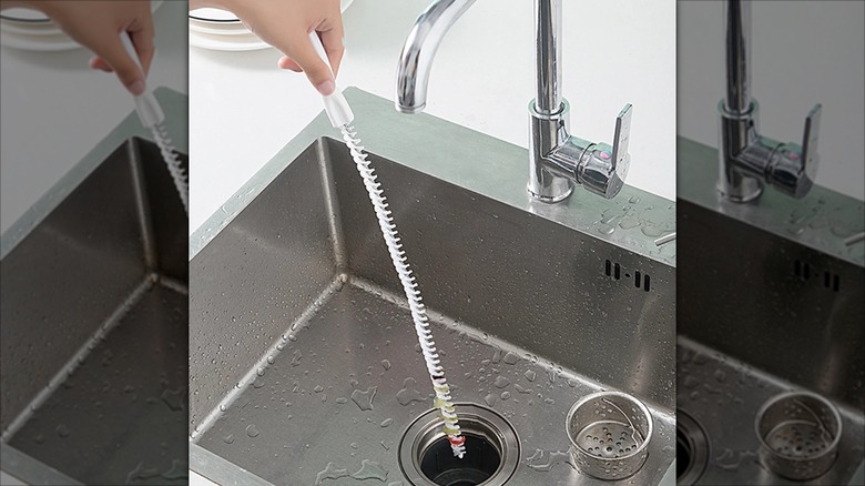 A person puts a white flexible bristle brush down a stainless steel sink drain
