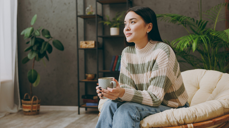 A woman sitting in a parlor cupping a hot drink in her hands.
