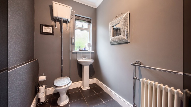The cistern of a high-tank toilet is installed close to the ceiling in a grey bathroom