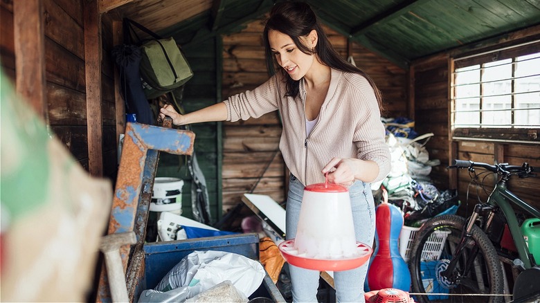 A smiling person stands in a cluttered garden shed and holds an empty hummingbird feeder