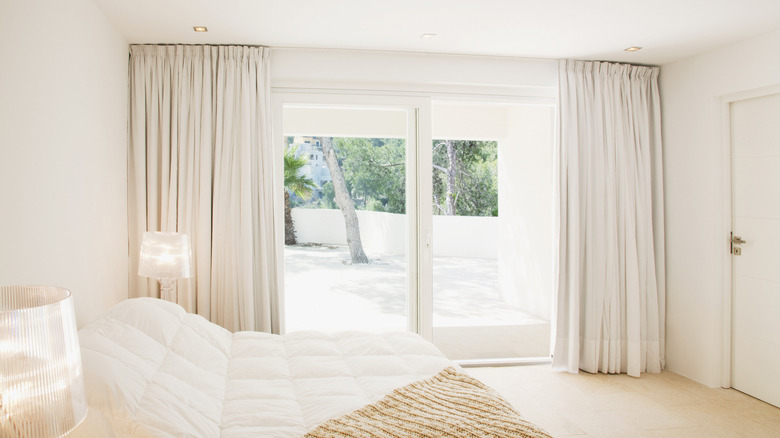 A white bedroom with white curtains pulled to the sides of a large glass sliding door