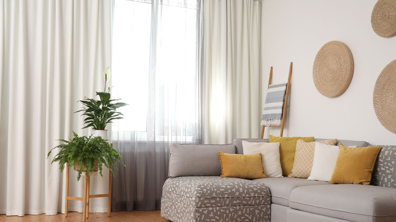 A grey couch with yellow throw pillows in front of a large, sunny window with open white curtains