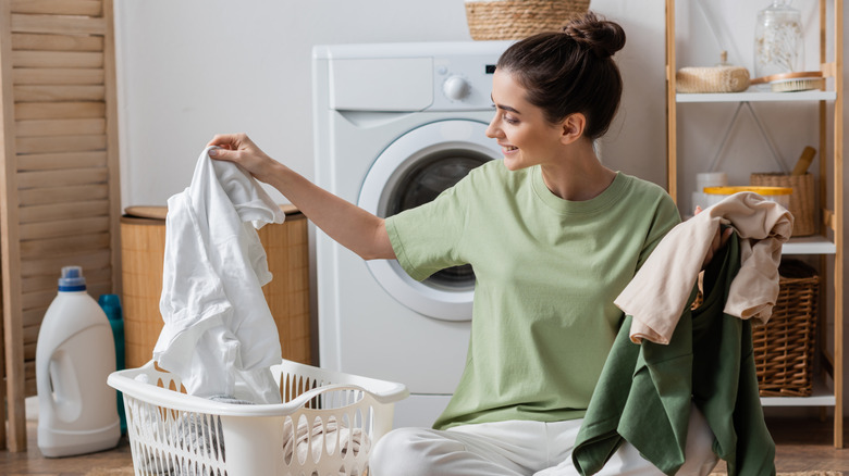 Woman wearing green shirt and white pants sits on the floor inside a laundry room to sort clean laundry