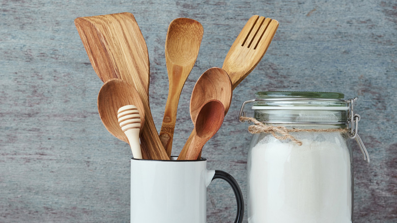 A white mug filled with wooden kitchen utensils next to a glass jar of sugar