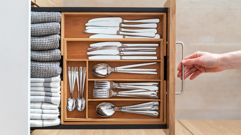 A hand is pulling open a drawer that contains silverware and small folded towels.