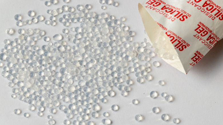 A white paper pouch labeled "Silica Gel" is open, with tiny clear gel beads spilling out onto a white surface.