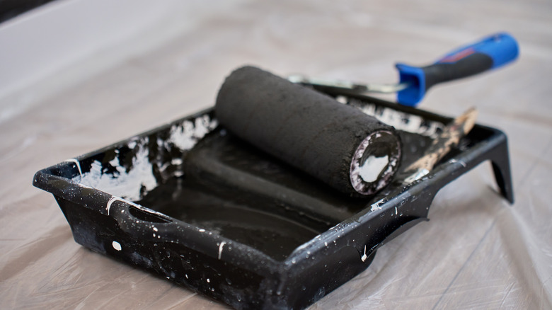 A paint roller with a blue handle laying in a tray of black paint with white residual paint on the sides.