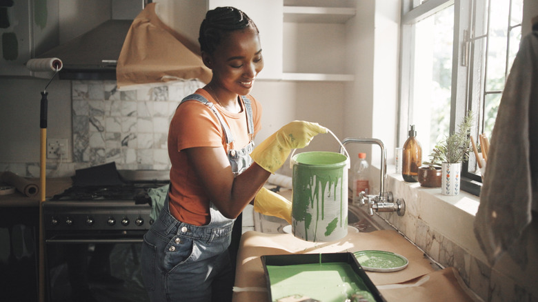 black woman in kitchen with can of olive green paint pouring into tray to get ready to paint