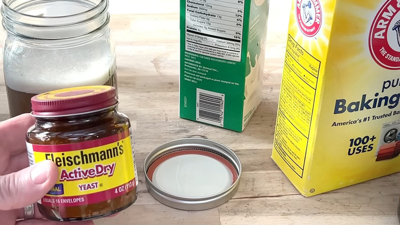 A hand holding a jar of active dry yeast with a fermented liquid in the background with other ingredients in background.