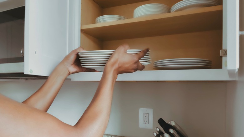 Person putting stack of white plates in white kitchen wall cabinet