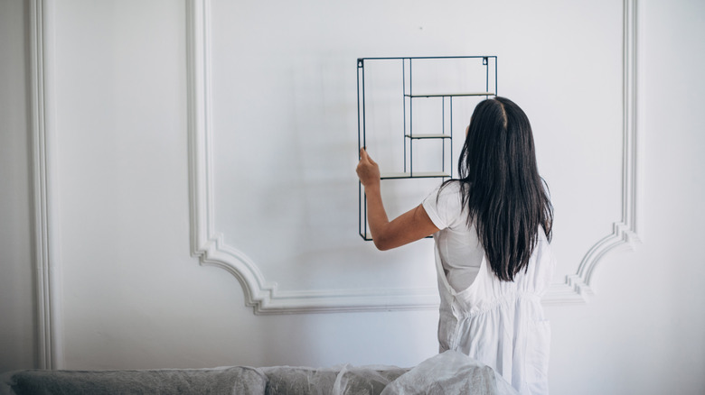 A long-haired woman hangs a small shelving unit on a wall in an entryway.