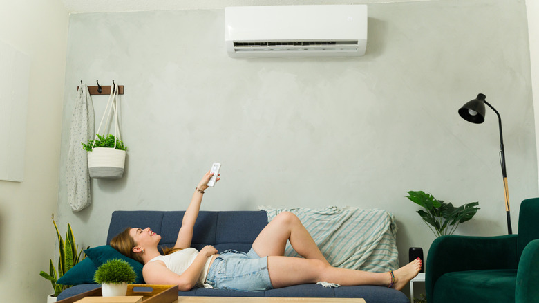Young woman lying on sofa using remote control, enjoying cool air from mini split ac unit during summer heat