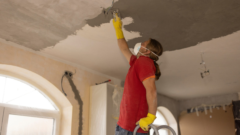 A man wearing a mask applies putty to a ceiling while standing on a ladder
