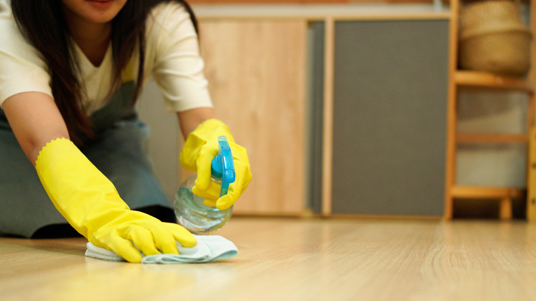 A person wearing yellow gloves uses a spray bottle and cloth to clean the floor.
