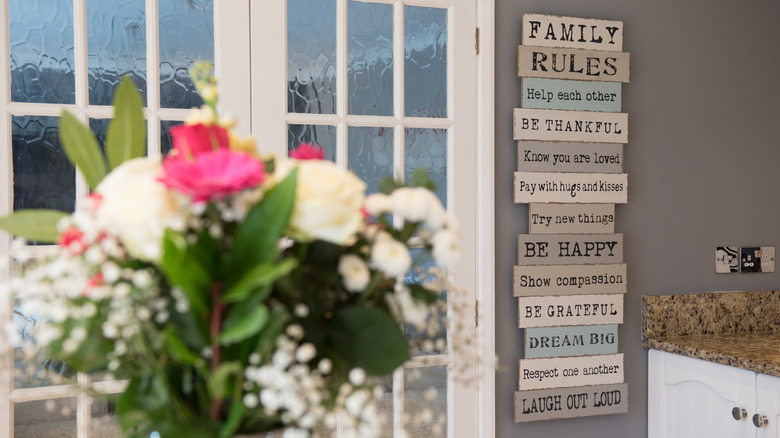 A general interior view of a sign with various sayings and quotations hanging on a wall in a kitchen with a vase of fresh cut flowers within a home.