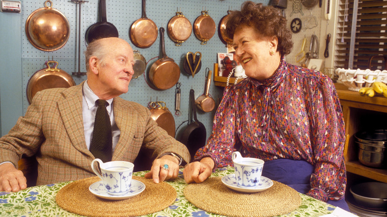 Julia Child and her husband sitting at a table, drinking from tea cups, and laughing