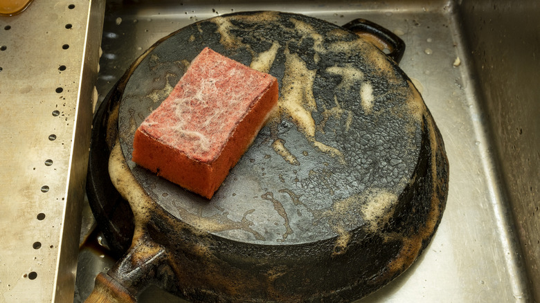 A cast iron pot turned upside down in the sink being scrubbed with a sponge and soap