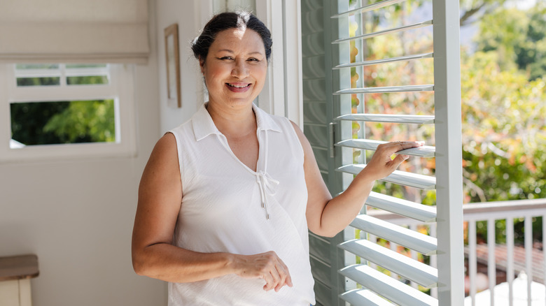 Smiling woman stands in sunshine in her home next to clean window shutters.