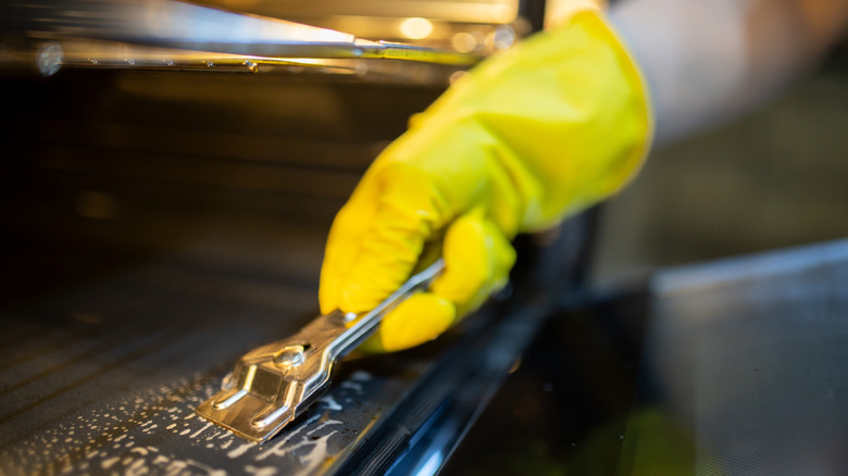 A yellow gloved hand scraping the inside of an oven using a metal scraper