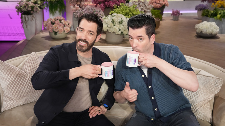 The Property Brothers' Jonathan and Drew Scott sitting on a sofa and drinking a hot beverage