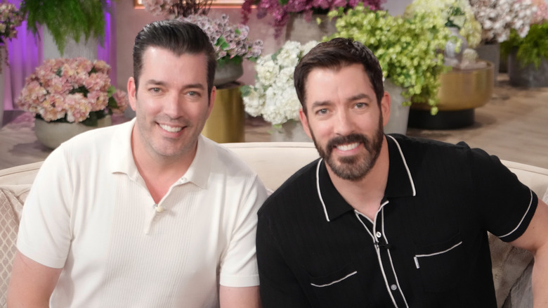 Jonathan and Drew Scott sitting side by side on a couch and smiling at the camera.