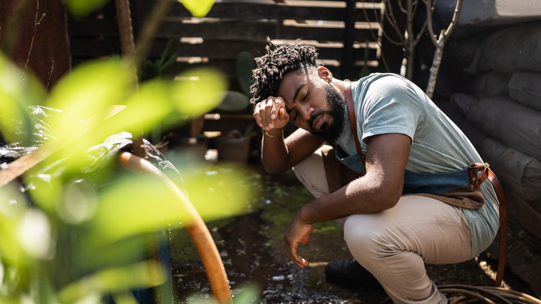 A young man in work clothes crouching in a garden area looking defeated
