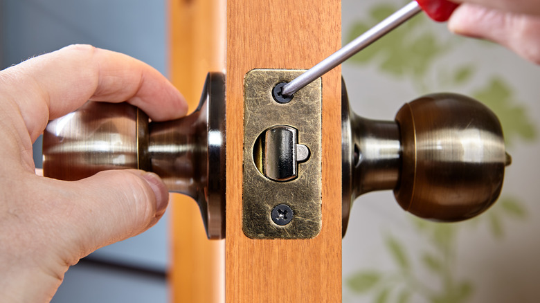 A doorknob is visible on both sides of a door and a person holds a screwdriver.