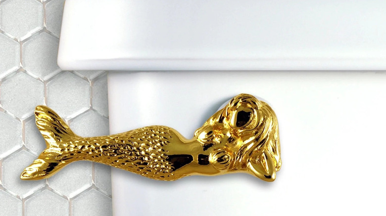 A shiny gold mermaid shaped toilet flusher is installed on a white toilet