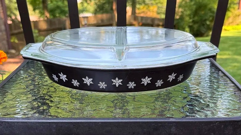 A Black Snowflake oval divided casserole dish with its clear lid sits on a glass table outside