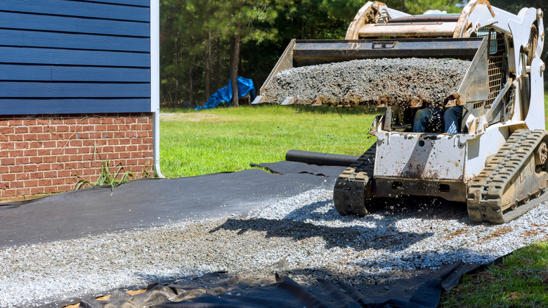 A person uses a loader machine to move gravel on top of a geotextile fabric for a driveway near a house