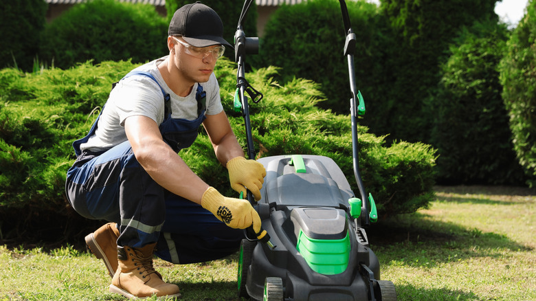A man in coveralls tightening a screw on his black and green lawn mower using a screwdriver