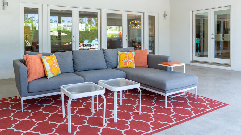 A concrete patio floor with a trellis-style red area rug and an outdoor gray sofa on top of it